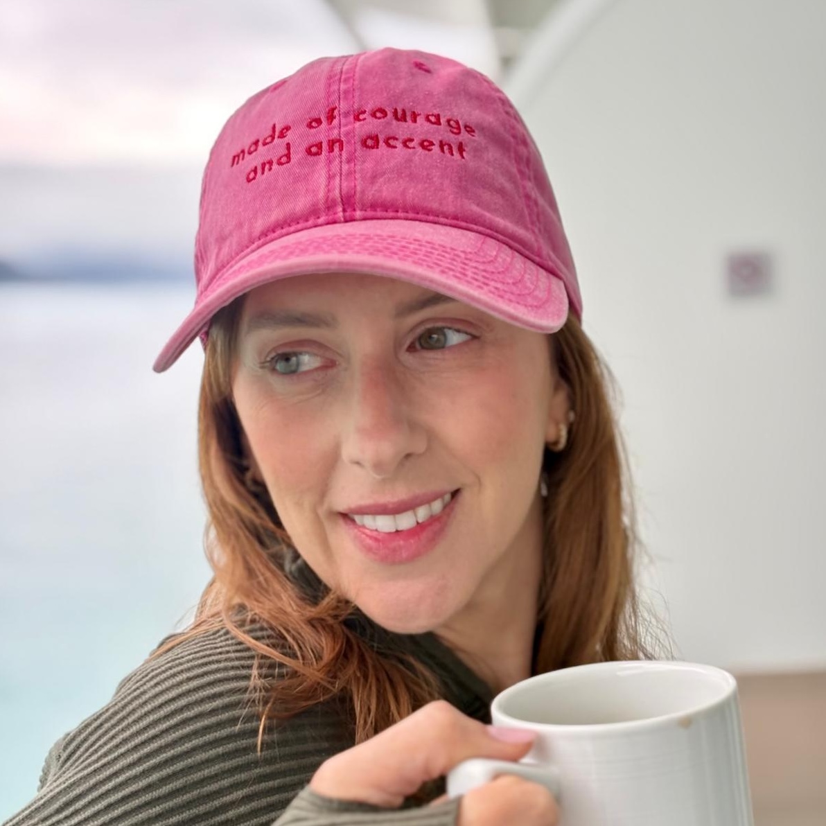 Hot pink acid-wash cap with tonal embroidery reading "made of courage and an accent" on a light oak table